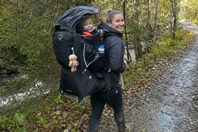 Mor går på en våt skogssti med et barn i en svart Osprey Poco Plus bæremeis med værbeskyttelse.